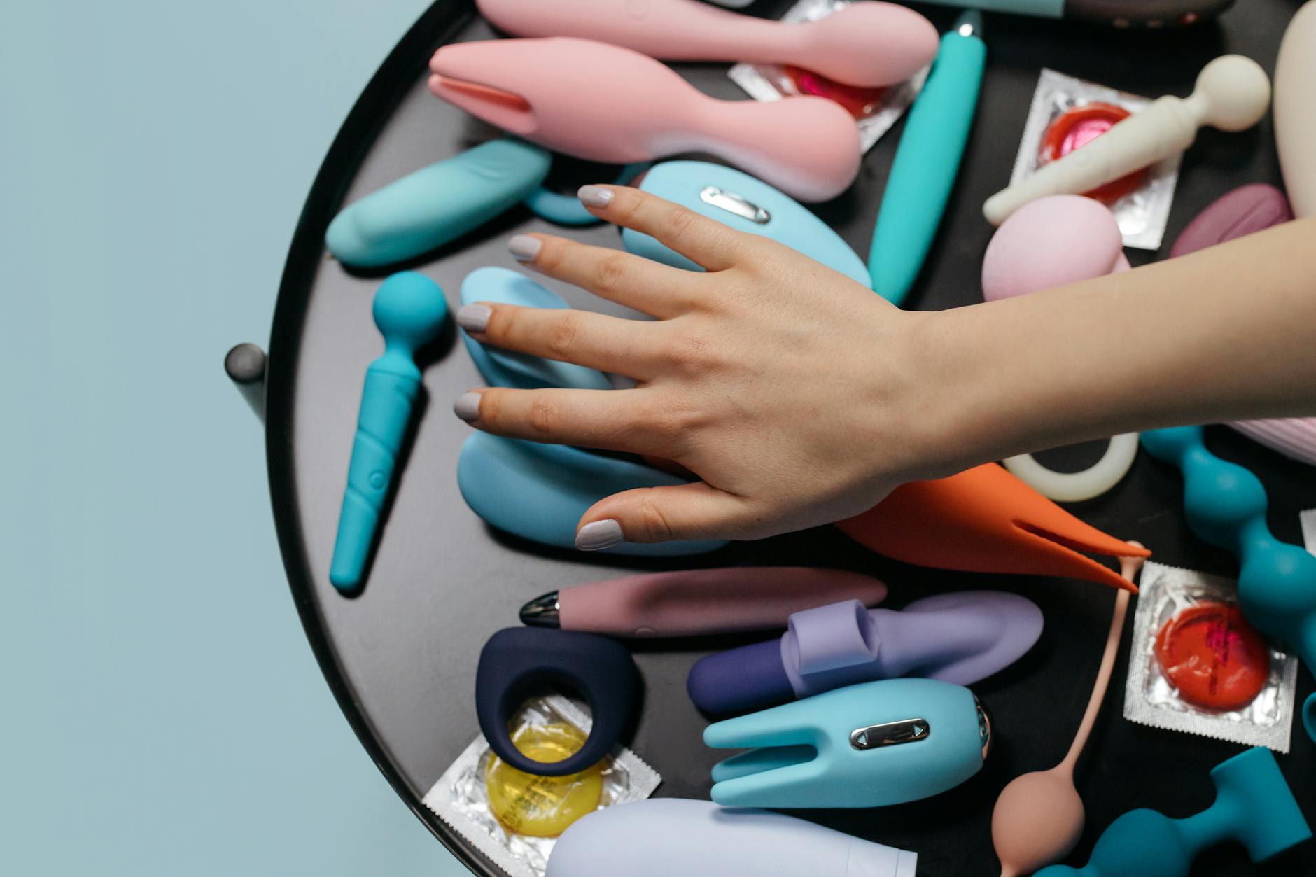 Hand reaching over an arrangement of colorful clitoral vibrators and adult toys on a table.